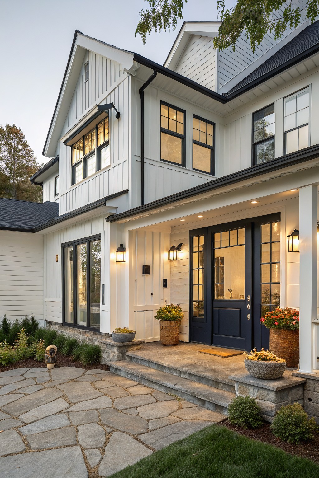 Two-story modern farmhouse exterior with white board-and-batten siding, navy blue glass-paneled double front doors, black window frames and trim, covered porch with white columns, stone steps and pathway, potted plants, and landscape lighting at dusk.