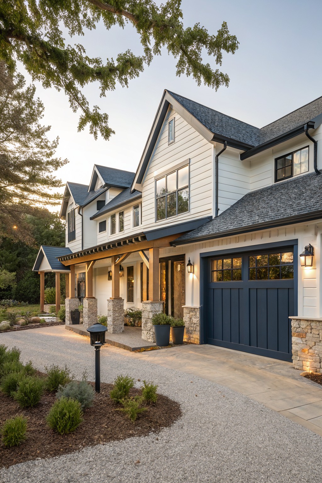 Side view of a modern farmhouse exterior featuring white board-and-batten siding, dark gray roof, navy blue two-car garage door, wooden covered porch with stone pillars, gravel driveway, low plantings, and mature trees at dusk.