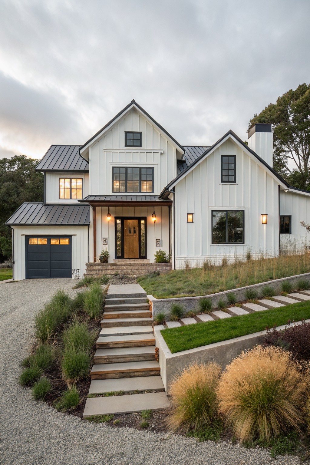 White modern farmhouse exterior with navy blue double garage doors, black-framed windows, dark metal roof, covered entry porch, and steps leading to gravel driveway amid native grasses.