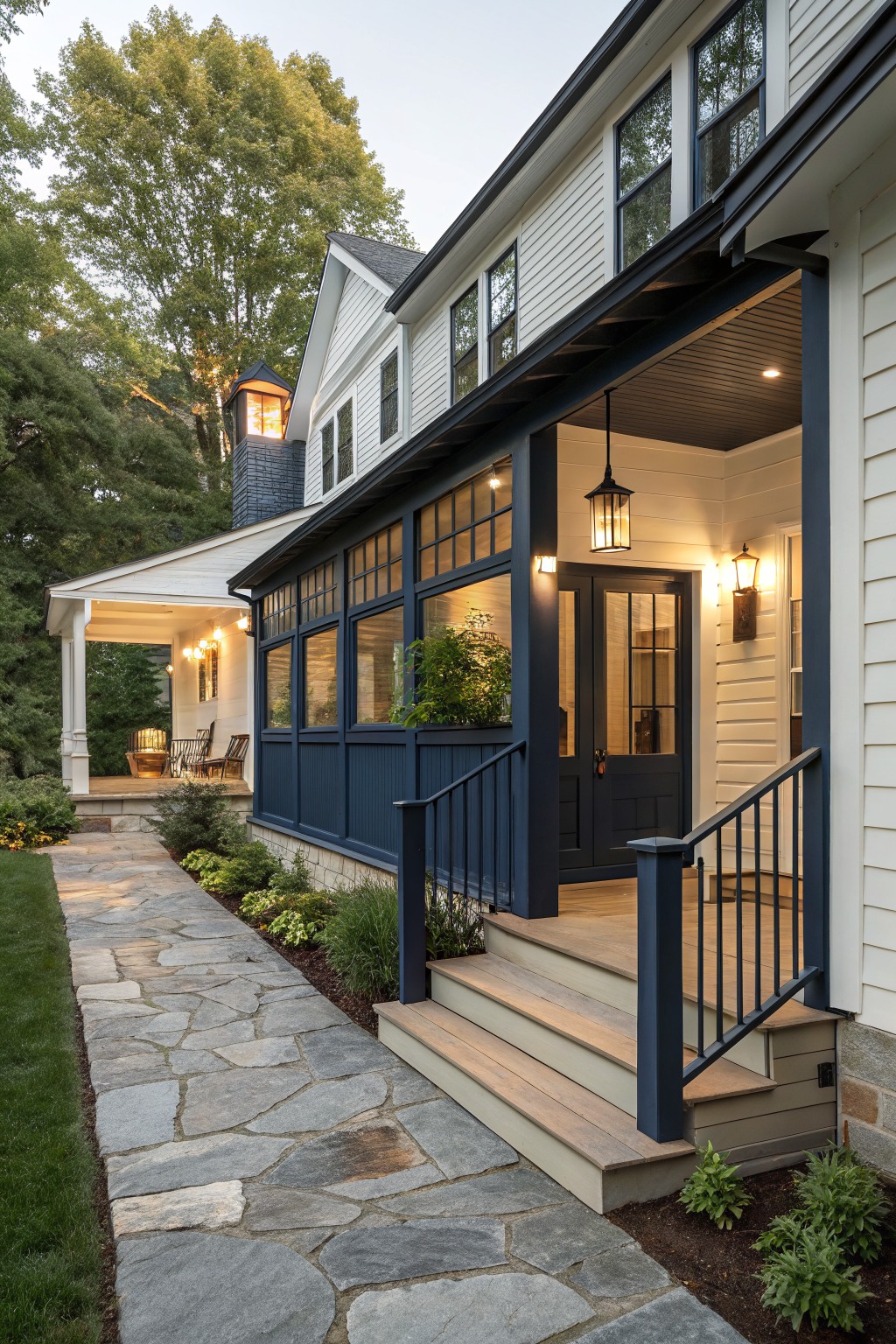 Side exterior of a white clapboard house featuring a navy blue enclosed porch with railings, dark door, pendant lights, stone pathway, landscaping, and trees at dusk.