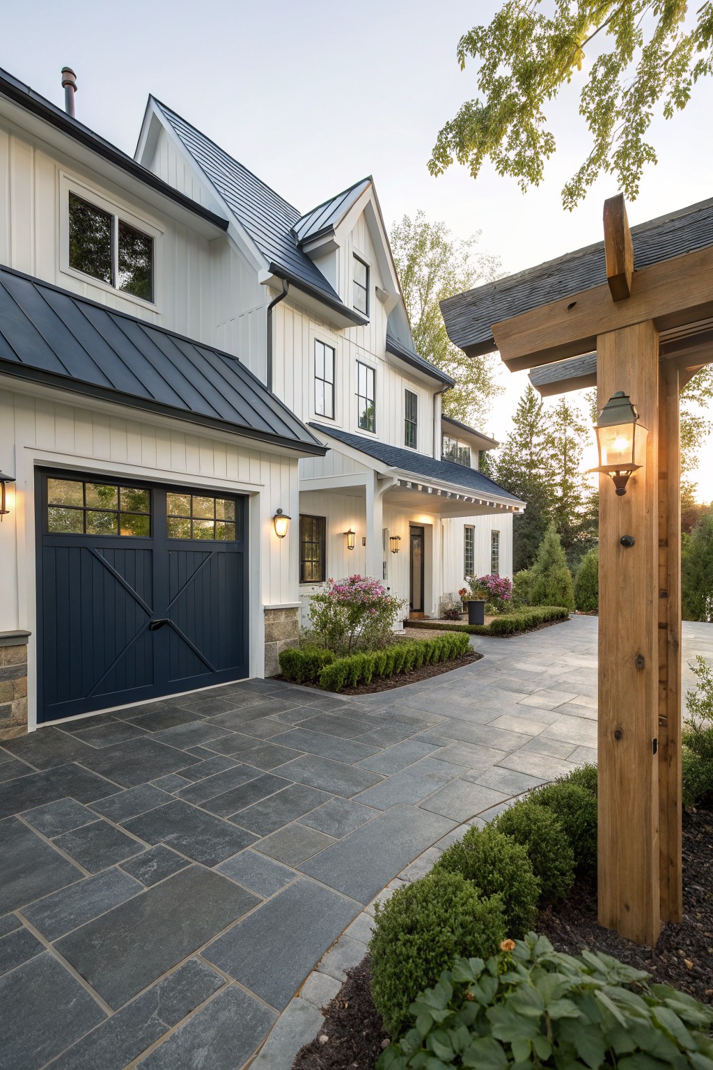 White two-story modern farmhouse exterior featuring navy blue standing-seam metal roof, navy blue wood garage door, black-framed windows, bluestone paver driveway, boxwood hedges, and wooden pergola post.