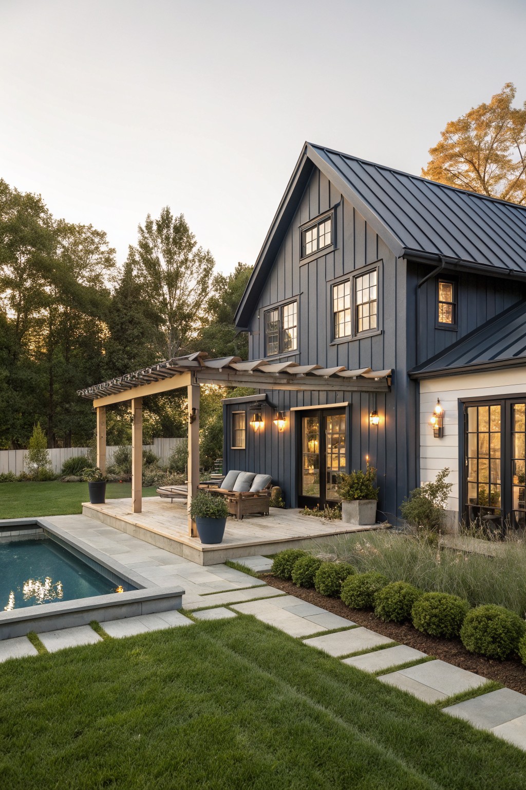Modern farmhouse exterior with navy blue board-and-batten siding, white trim on one side, standing seam metal roof, pergola over wooden deck, inground pool, bluestone pavers, boxwood shrubs, and lawn at dusk.