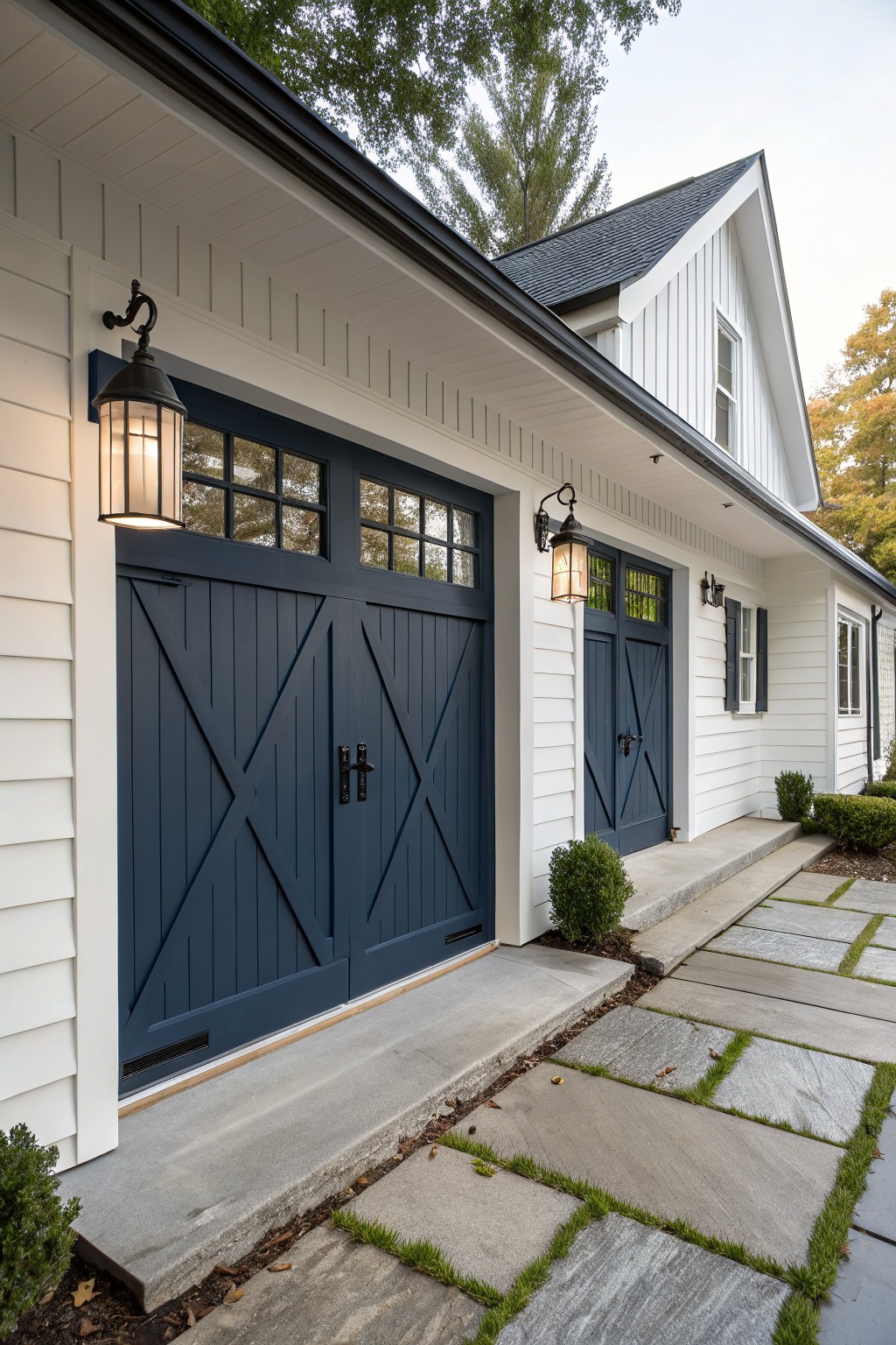 White vertical board-and-batten house exterior featuring two navy blue X-braced double garage doors with glass panels, flanked by black lanterns, a concrete apron, bluestone paver walkway edged in grass, and low boxwood shrubs.