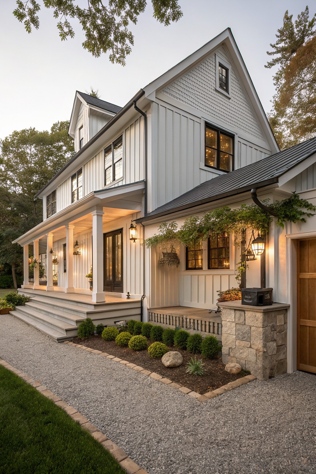 Two-story white board-and-batten sided farmhouse exterior with dark metal roof, wraparound porch supported by columns, wood garage door with stone pillars, gravel pathway, shrubs, and landscape lighting at dusk.