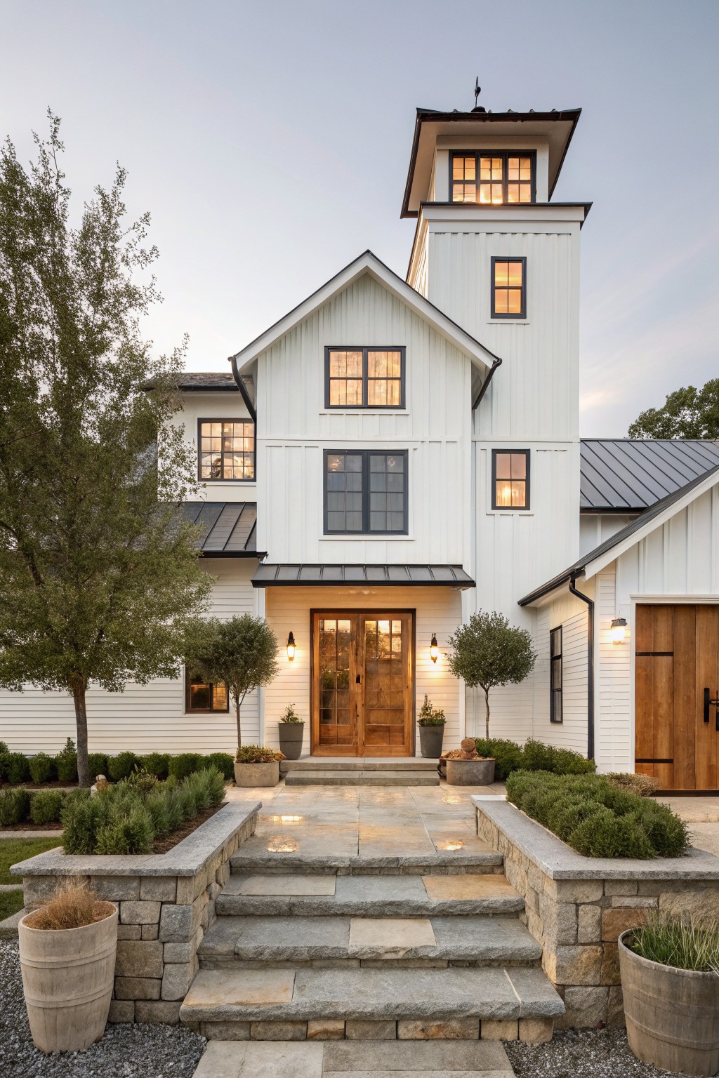 Front view of a two-story white shiplap modern farmhouse at dusk with a tall tower, black-framed windows, double wood entry doors, wood garage door, metal roof, stone steps, boxwood hedges, and potted plants.