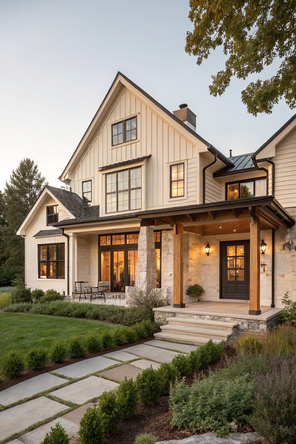 Two-story modern farmhouse exterior with light board-and-batten siding, covered front porch on stone base supported by dark timber posts, large windows, dark wood entry door, and landscaped yard with shrubs, grass, and stone path.