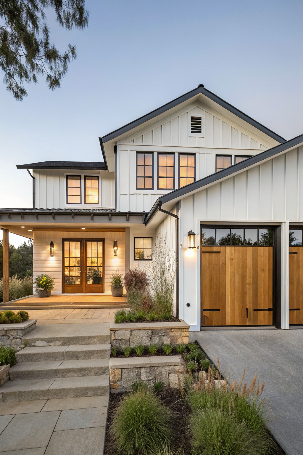 A modern farmhouse house exterior featuring white shiplap siding, black metal roof and trim, paired wooden garage doors, a covered front porch with double glass doors, stone steps, and low plantings along the driveway at dusk.