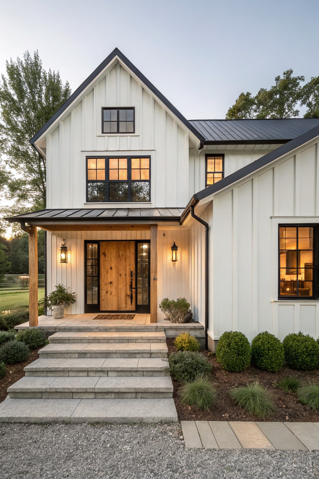 White shiplap modern farmhouse house with gabled roof, black metal roofing, wooden double front doors, covered porch with lanterns, stone steps, gravel driveway, and shrubs at dusk.