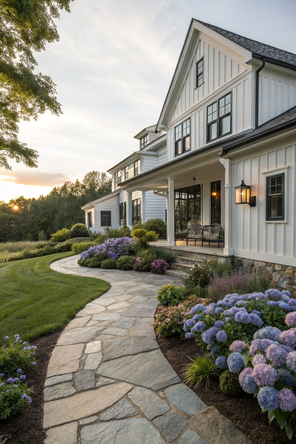 White shiplap farmhouse with wraparound porch and stone foundation, viewed from the yard with a curved flagstone path lined by purple hydrangea bushes and other perennials leading to porch steps, green lawn and trees in background at sunset.