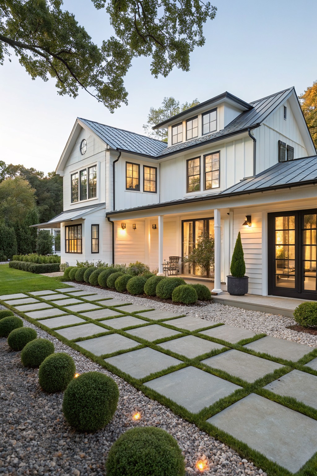 White modern farmhouse exterior with metal roof, black-framed windows, and covered porch approached by a grid of large rectangular concrete pavers with grass joints, flanked by spherical boxwood shrubs and gravel edging in a front yard at dusk.