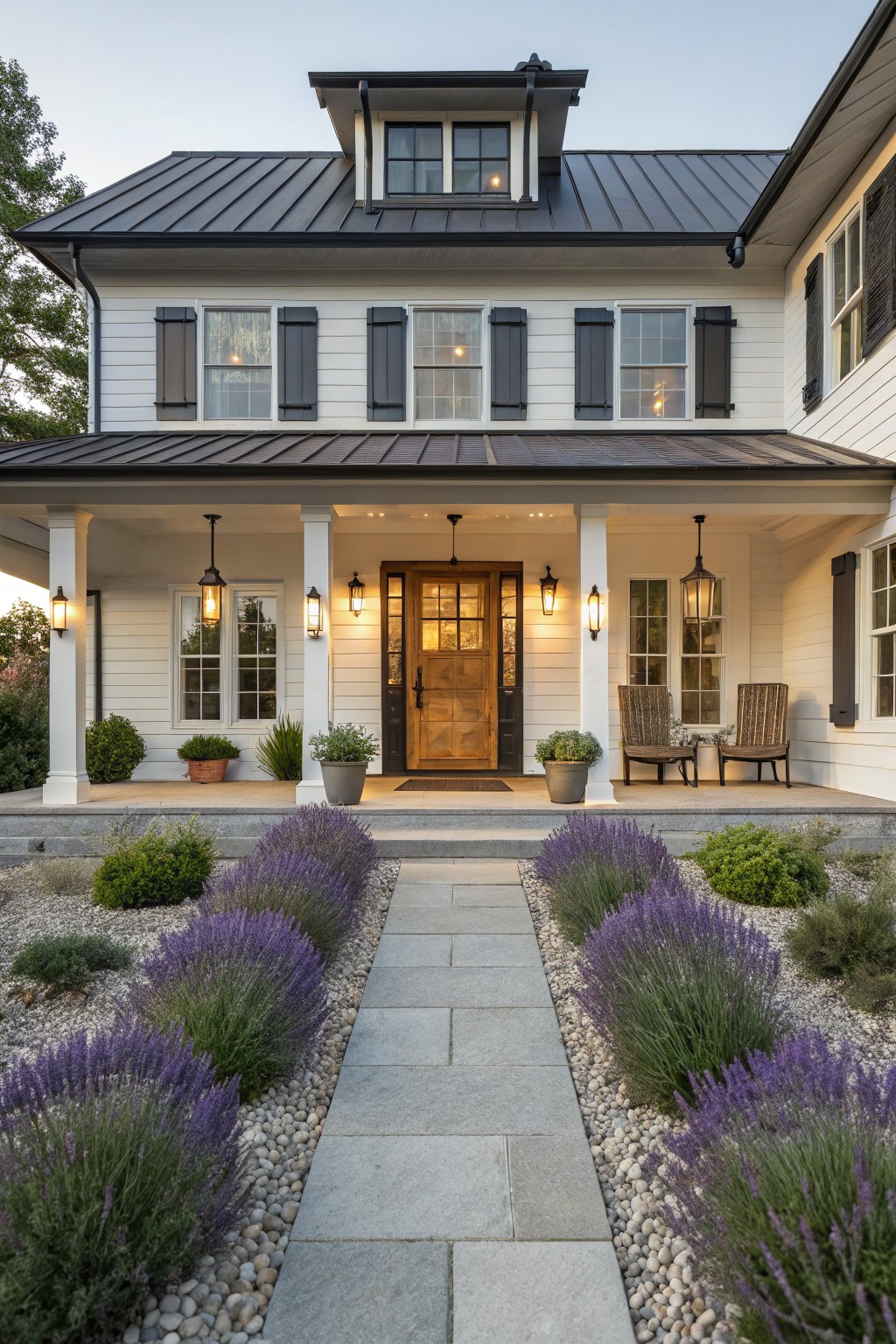 White clapboard house with black metal roof, shutters, and porch columns, wooden front door, flanked by potted plants and chairs, with gray stone pathway lined by lavender bushes and gravel mulch in the front yard.
