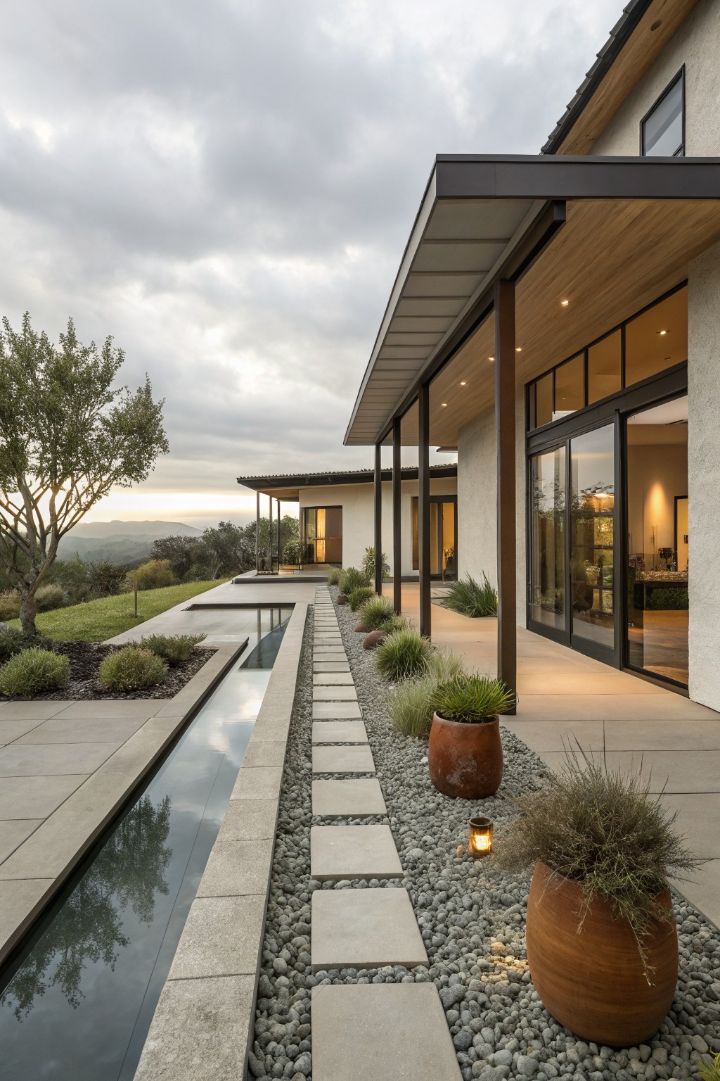 Side view of a modern stucco house with covered wooden terrace and sliding glass doors, featuring a linear concrete water channel parallel to a gravel path with square stone stepping stones, flanked by potted grasses and hillside landscape.