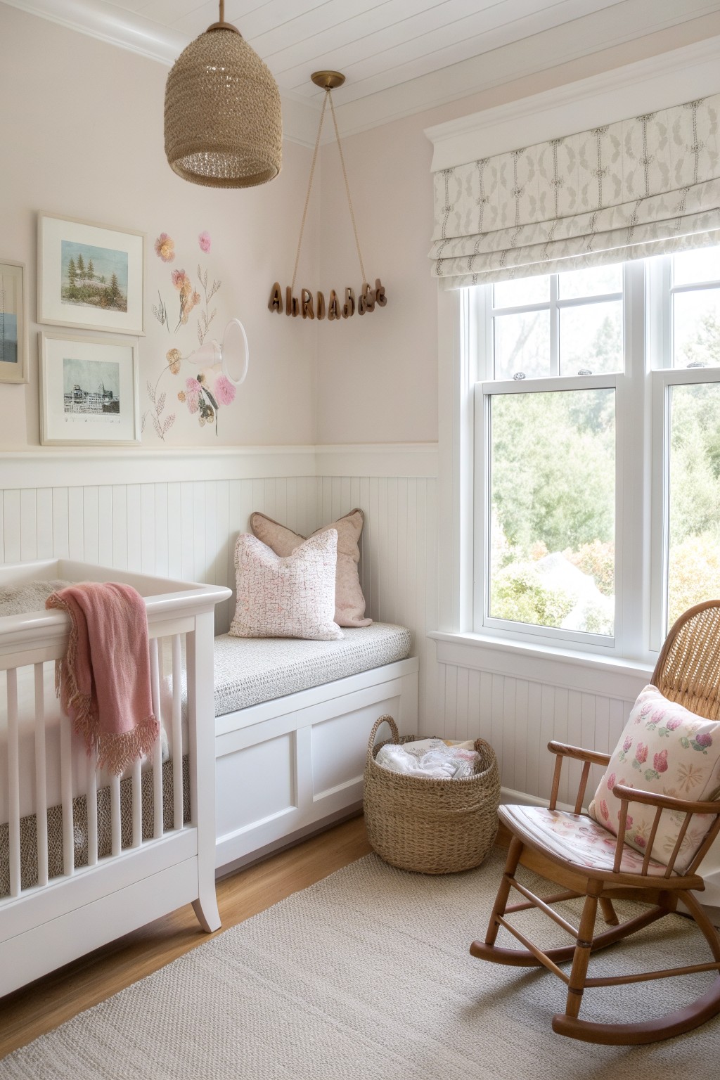 Nursery room with white beadboard wainscoting on lower walls, white crib draped in pink blanket, built-in window seat with pink pillows, wicker basket, wooden rocking chair with floral cushion, and large window with neutral roman shade overlooking trees.