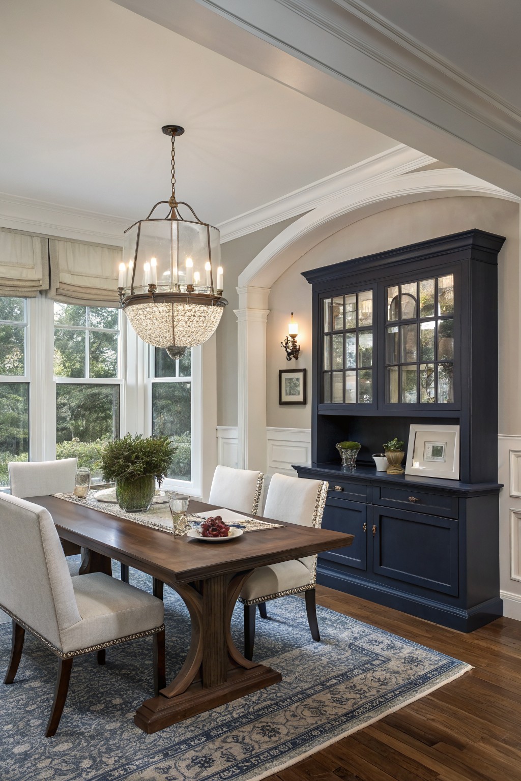 Dining room interior with rectangular wooden table, white upholstered chairs, navy blue glass-front cabinet, crystal chandelier, beige walls, hardwood floors, and large multipane windows with white trim.