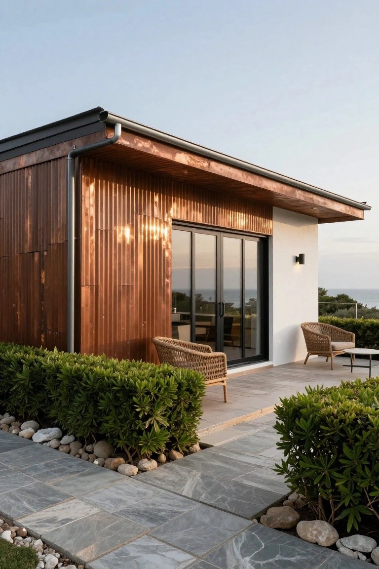 Modern single-story house exterior featuring vertical corrugated copper metal cladding on the side wall, black-framed glass sliding doors opening to a wooden deck with wicker chairs and a side table, white stucco wall, boxwood hedges, pebble mulch, and gray slate pavers, with ocean view in background.