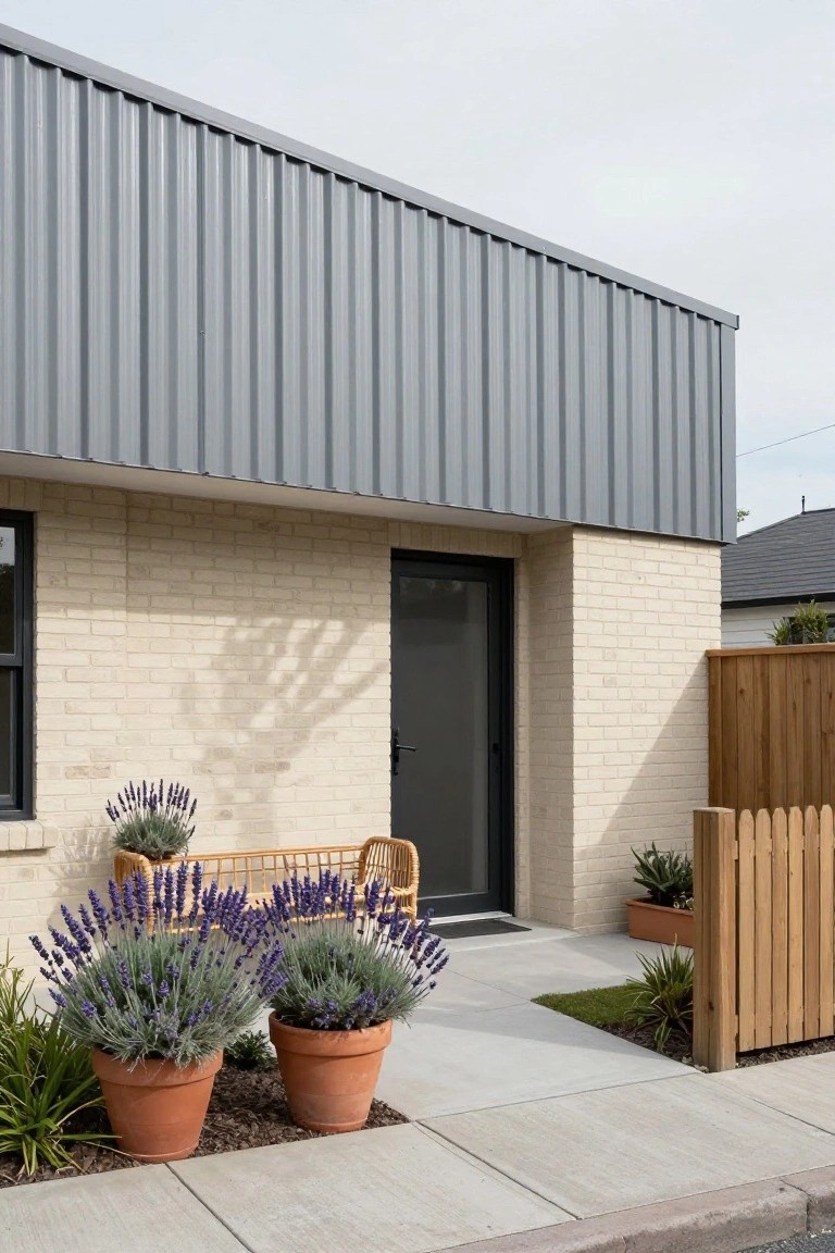 Front exterior of a modern single-story house with gray corrugated metal cladding on the upper wall and roof, light beige brick lower walls, black front door with sidelight, potted lavender plants, rattan bench, and wooden picket fence beside a concrete path.
