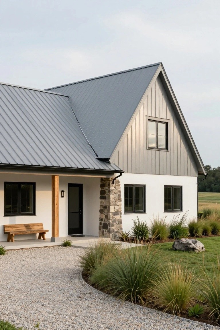 House exterior with gray standing seam metal gabled roof, white plaster walls, gray wood cladding on gable, stone pillar accents, black windows and door, wooden bench on porch, gravel path edged by ornamental grasses and rocks.