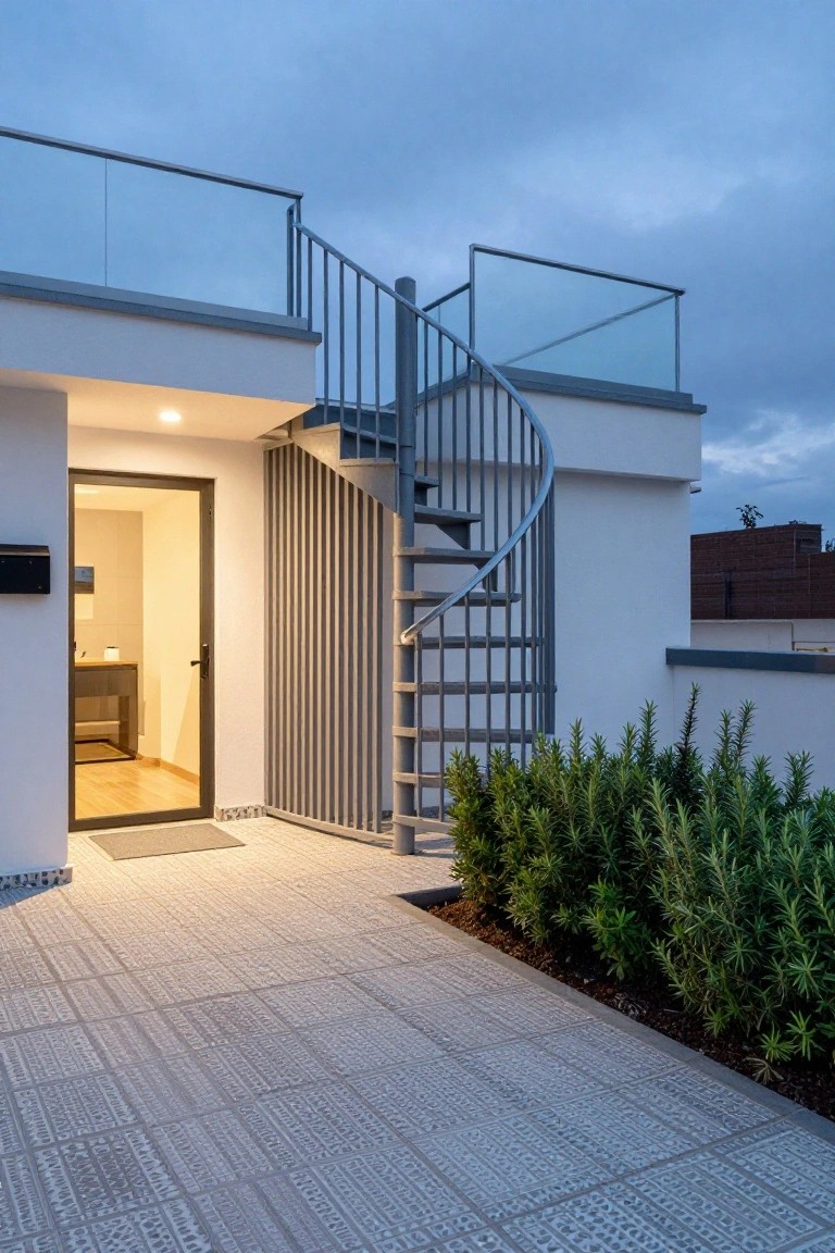 White modern house exterior featuring a glass front door, a gray metal spiral staircase with glass railing leading to an upper terrace, a tiled patio, and low rosemary bushes along the edge at dusk.