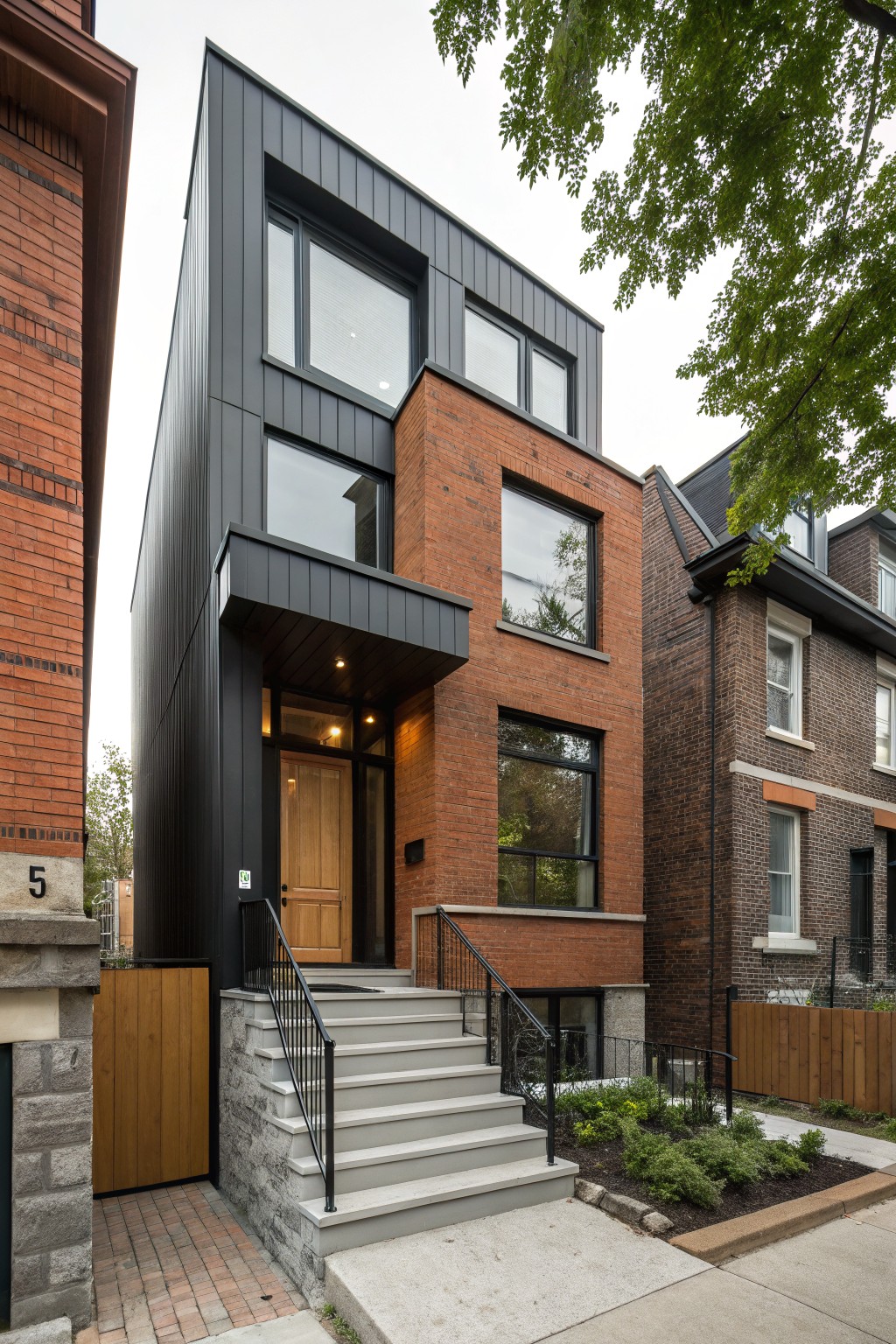 Three-story row house with black metal siding on upper sections and front corner, red brick base and side wall, large rectangular windows, wooden front door, metal railing on concrete steps, between traditional brick neighbors on a paved street with small front planting bed.
