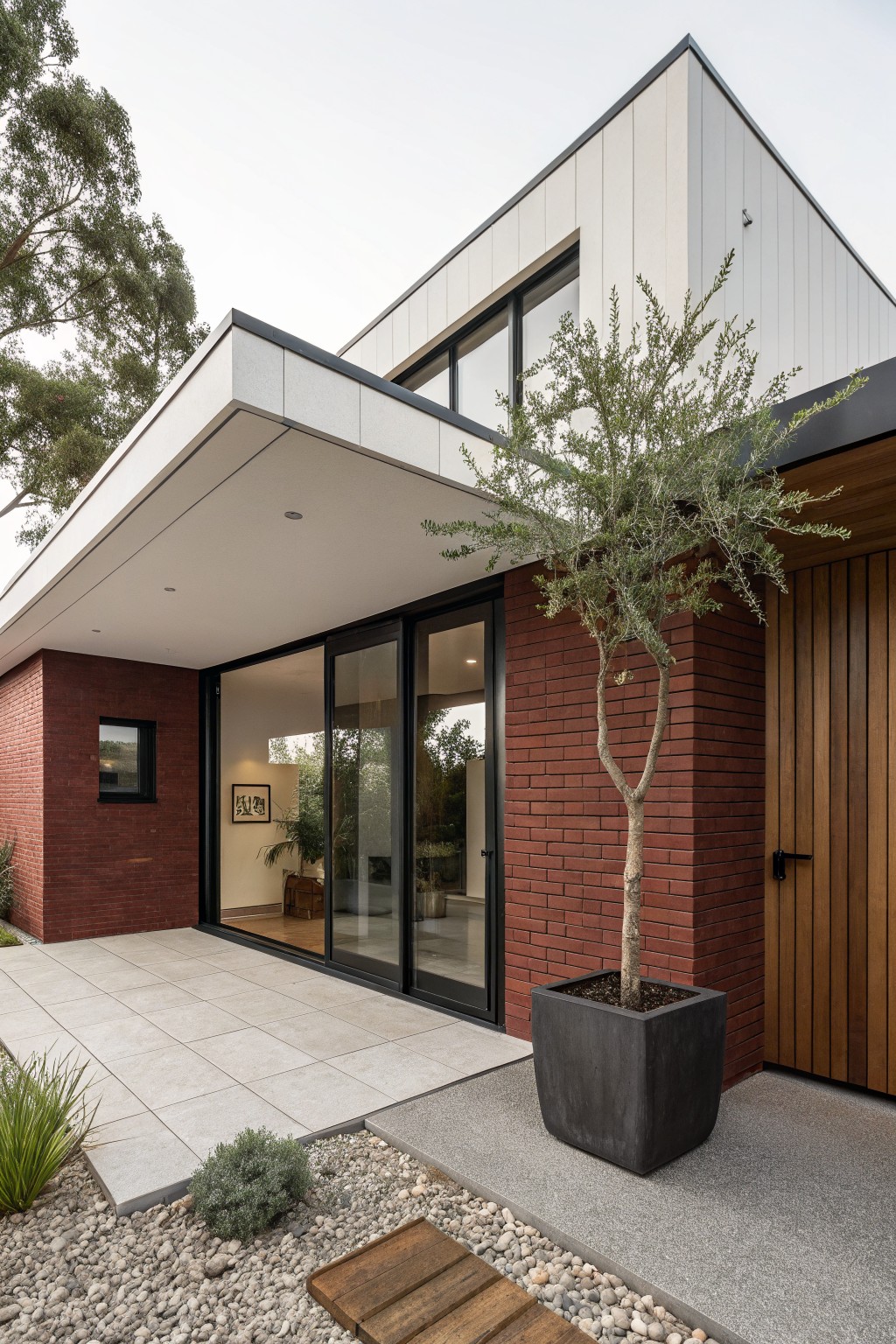 Modern house exterior with red brick lower walls, white vertical siding on upper sections, black-framed sliding glass doors and windows opening to a tiled patio, wooden garage door, potted olive tree beside brick, and gravel landscaping with low plants.