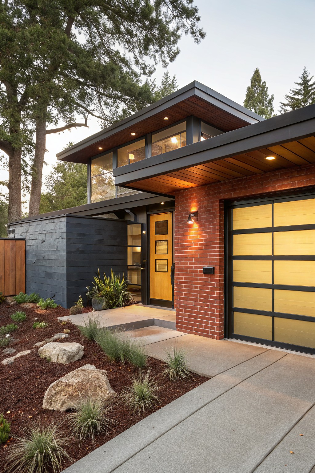 Modern two-story house exterior with red brick on the garage side, dark slate siding on angular volumes, wood overhangs and trim, yellow wood entry door, glass-paneled garage door, and gravel front yard with grasses and rocks.