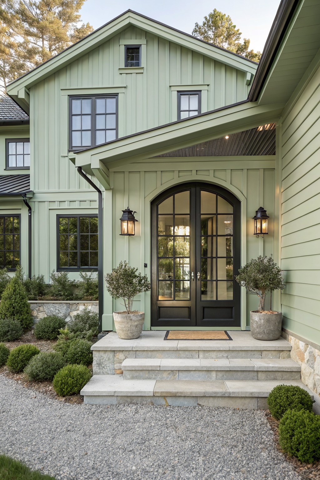 Sage green vertical board-and-batten sided house exterior featuring tall arched black double front doors with glass panels, flanked by black lanterns and potted olive trees, leading to stone steps amid gravel path and boxwood shrubs.