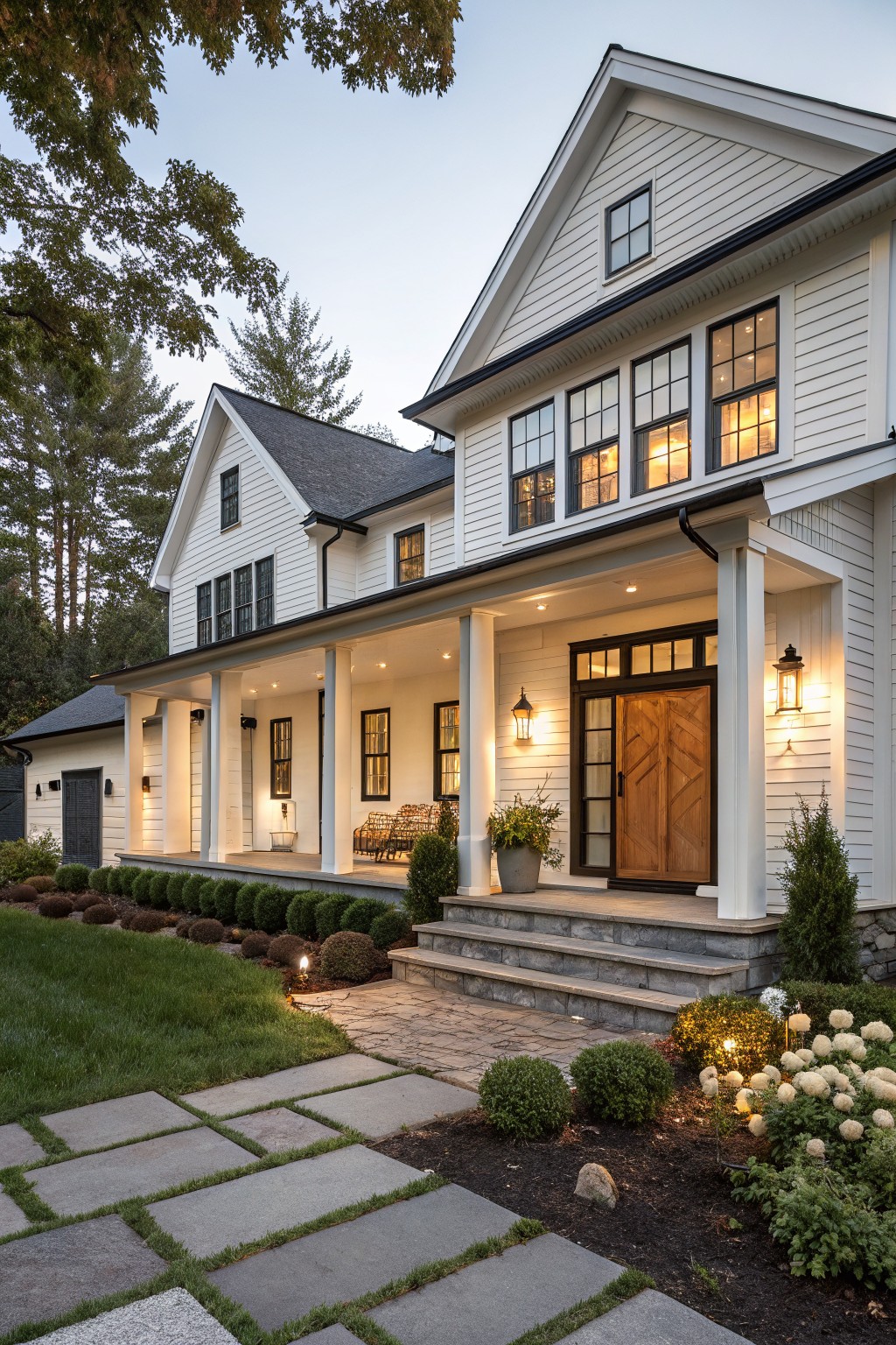 White board-and-batten farmhouse exterior with black window and door frames, chevron-patterned wooden front door under a covered porch with columns, stone steps, landscaped shrubs, and a bluestone path at dusk.