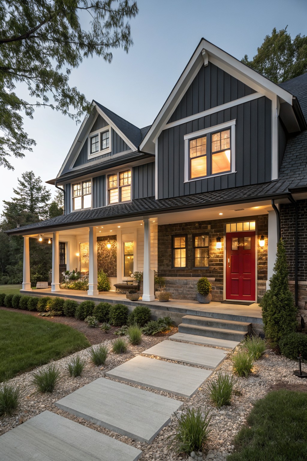 Two-story transitional farmhouse exterior featuring dark gray board-and-batten siding, white trim, black-framed windows, red front door, covered porch with columns, stone base, and concrete stepping stones leading to the entry amid low shrubs and gravel at dusk.