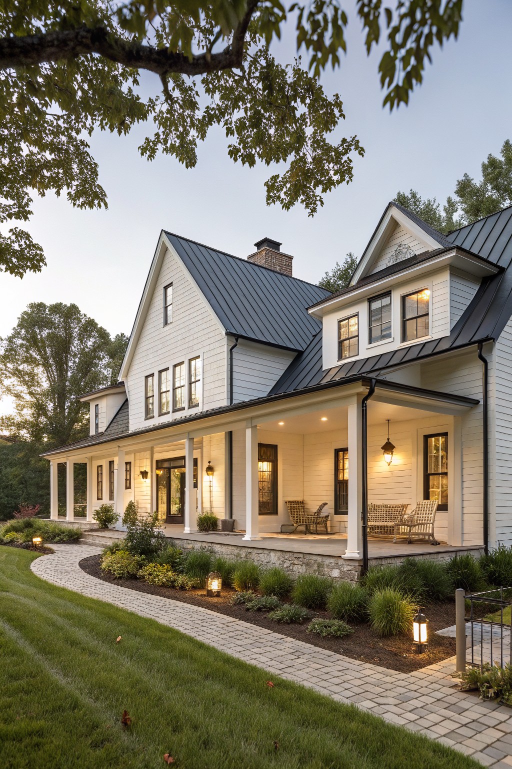 White board-and-batten farmhouse with dark standing-seam metal roof, wraparound porch with lanterns, and curved brick pathway through landscaped front yard at dusk.