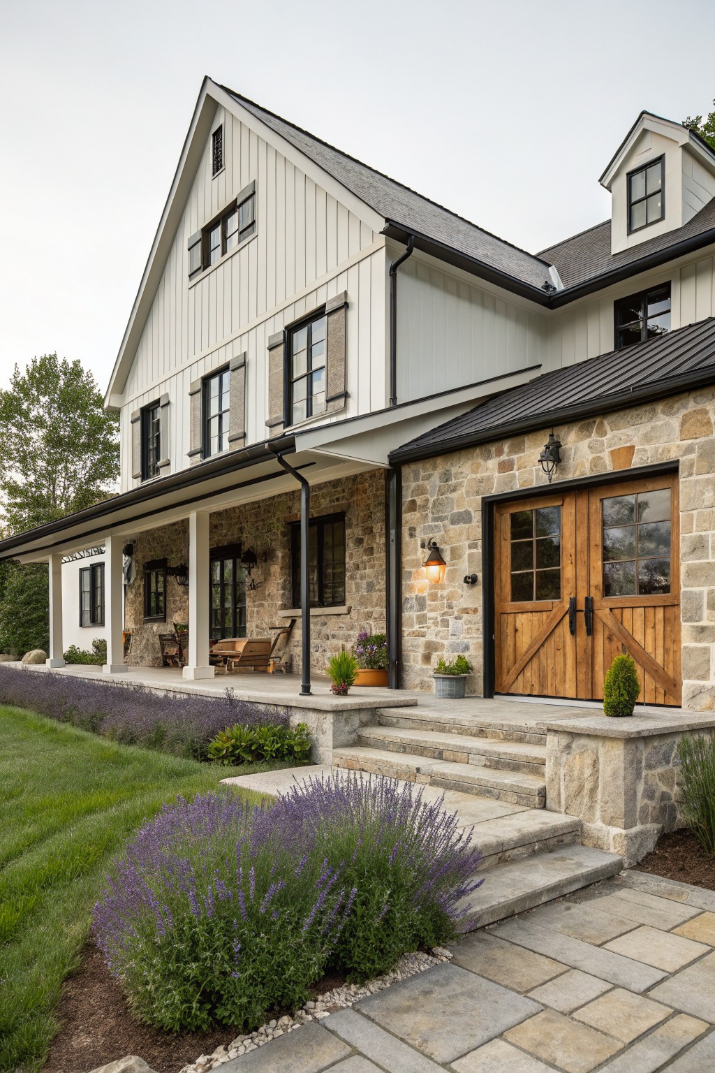 Two-story transitional farmhouse exterior with white board-and-batten siding, black window frames and shutters, metal roof, stone base and garage with wooden doors, wraparound porch, stone steps, and lavender plantings.