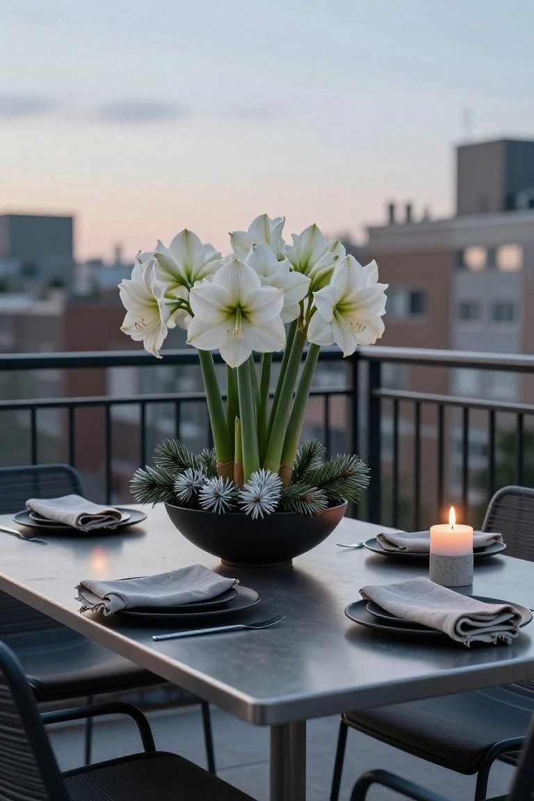 Black bowl on a metal outdoor dining table holding white amaryllis flowers with green stems, pine branches, and white spheres, next to a lit gray candle, with black plates, linen napkins, and chairs on a balcony overlooking city buildings at dusk.