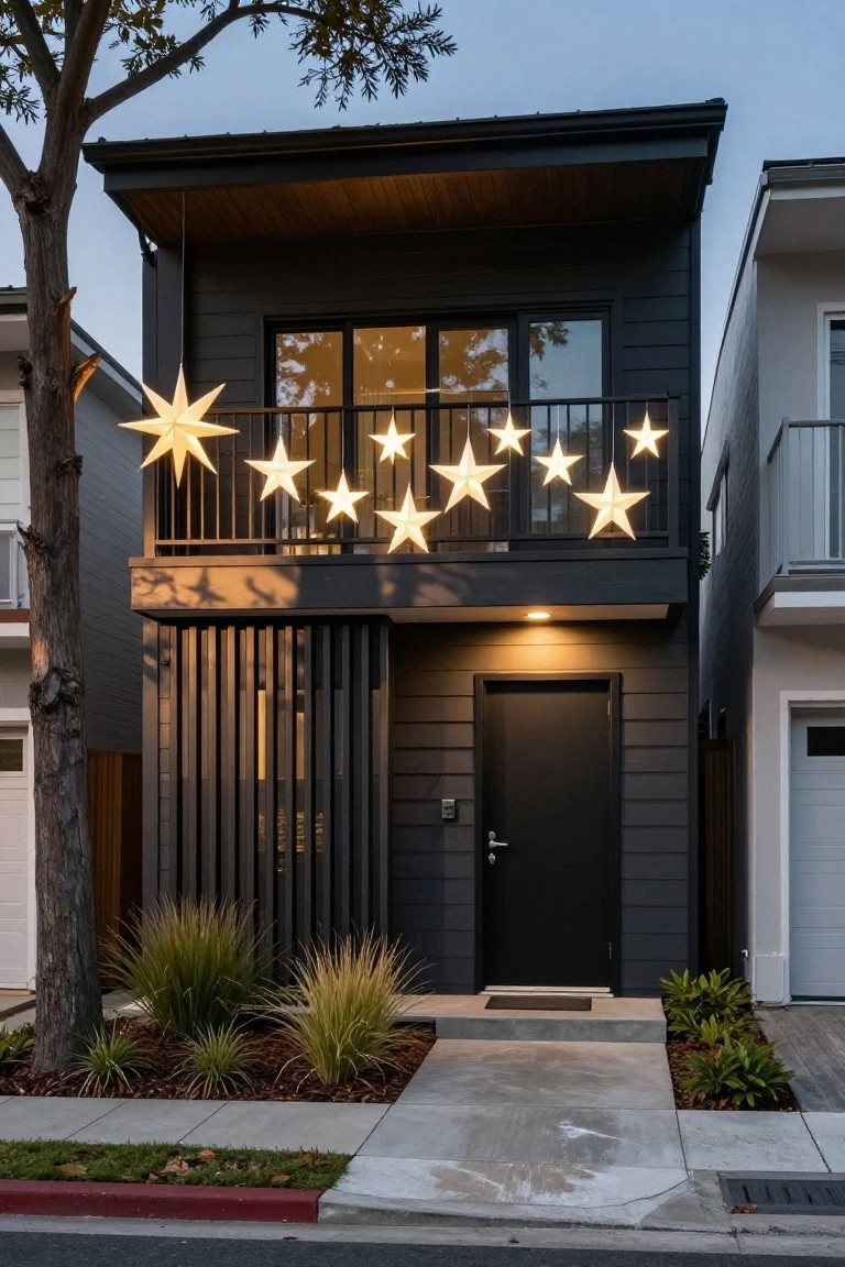 Modern two-story black house exterior at dusk with multiple large white paper star lights hanging from the second-floor balcony railing, dark wood accents, slatted entry screen, front landscaping, and neighboring homes visible.