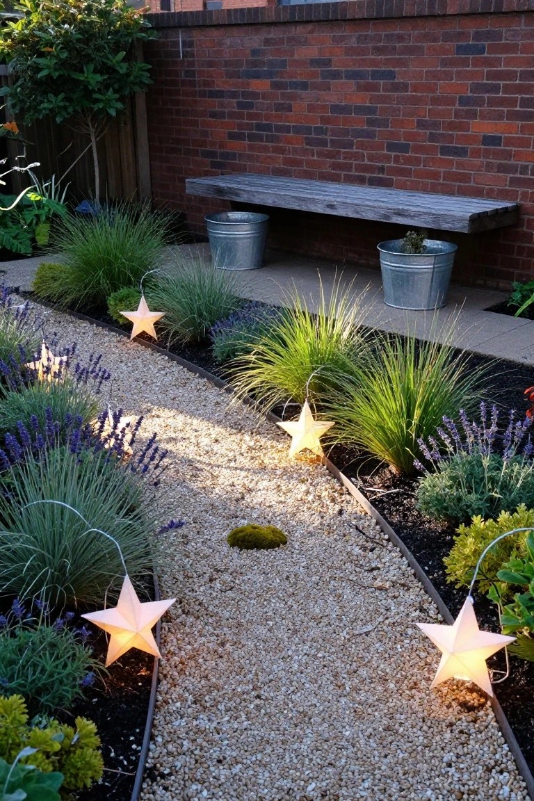 Winding gravel garden path edged with lavender, ornamental grasses, and metal buckets containing plants, lit by several hanging white paper star lanterns, with a wooden bench against a brick wall in the background.