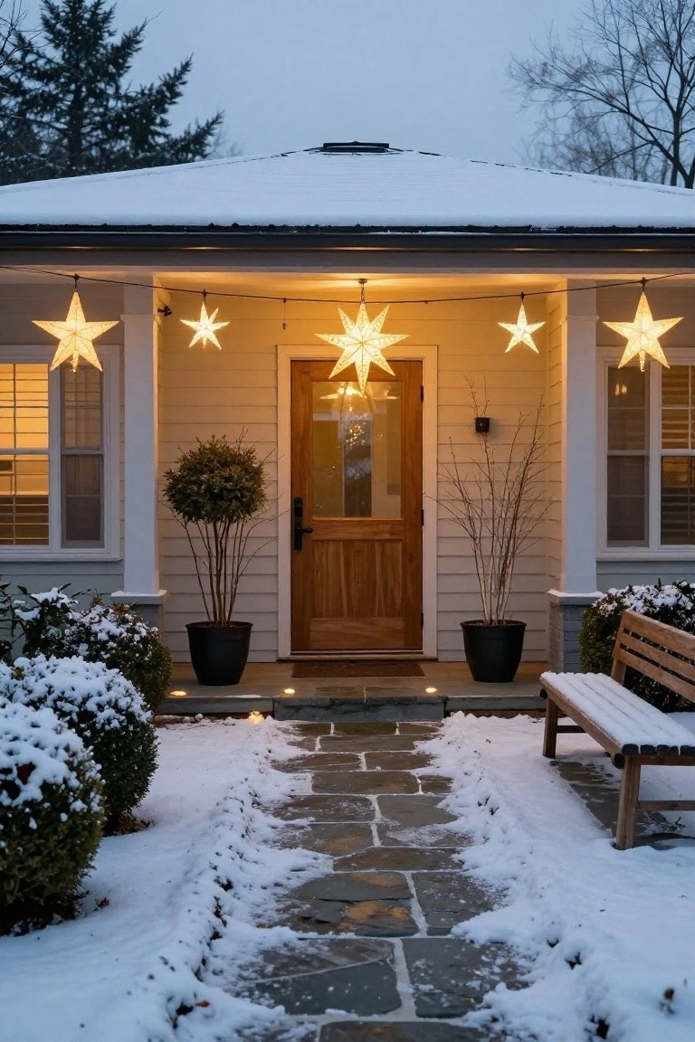 Snow-covered front porch of a light beige house with large glowing paper star lanterns hanging from the ceiling above a wooden door, flanked by potted shrubs and a stone pathway leading to the entry.