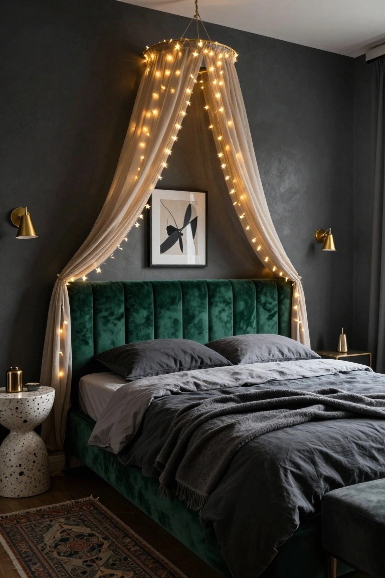 Bedroom interior with emerald green velvet tufted headboard and bed dressed in gray linens, framed by beige sheer canopy draped with white fairy lights from a gold hoop, dark textured walls, gold sconces, and speckled white bedside table.