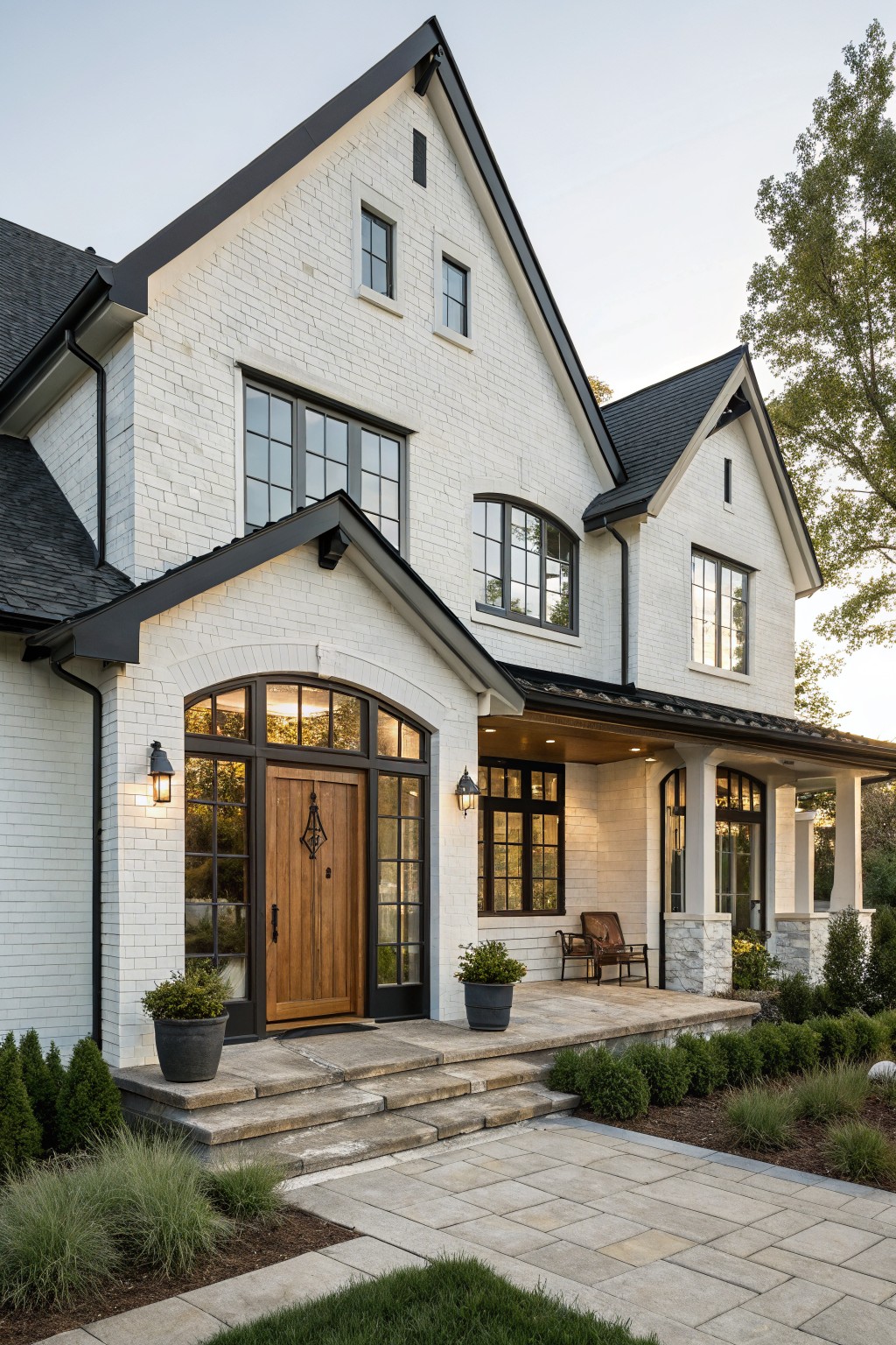 Two-story white brick house exterior with black window frames, dark gabled roof, arched wooden entry door with transom, covered front porch with chairs, stone steps, and low plantings along a paver path.