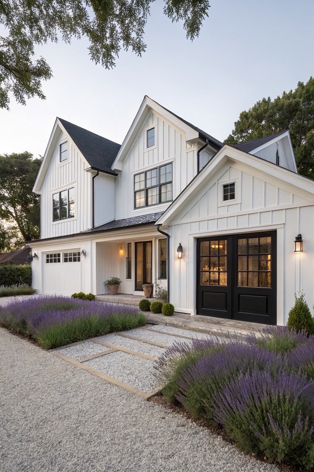 White board-and-batten farmhouse exterior with black-framed windows, double garage doors, black entry doors, lantern lights, gravel driveway, lavender plant borders, and stone path.