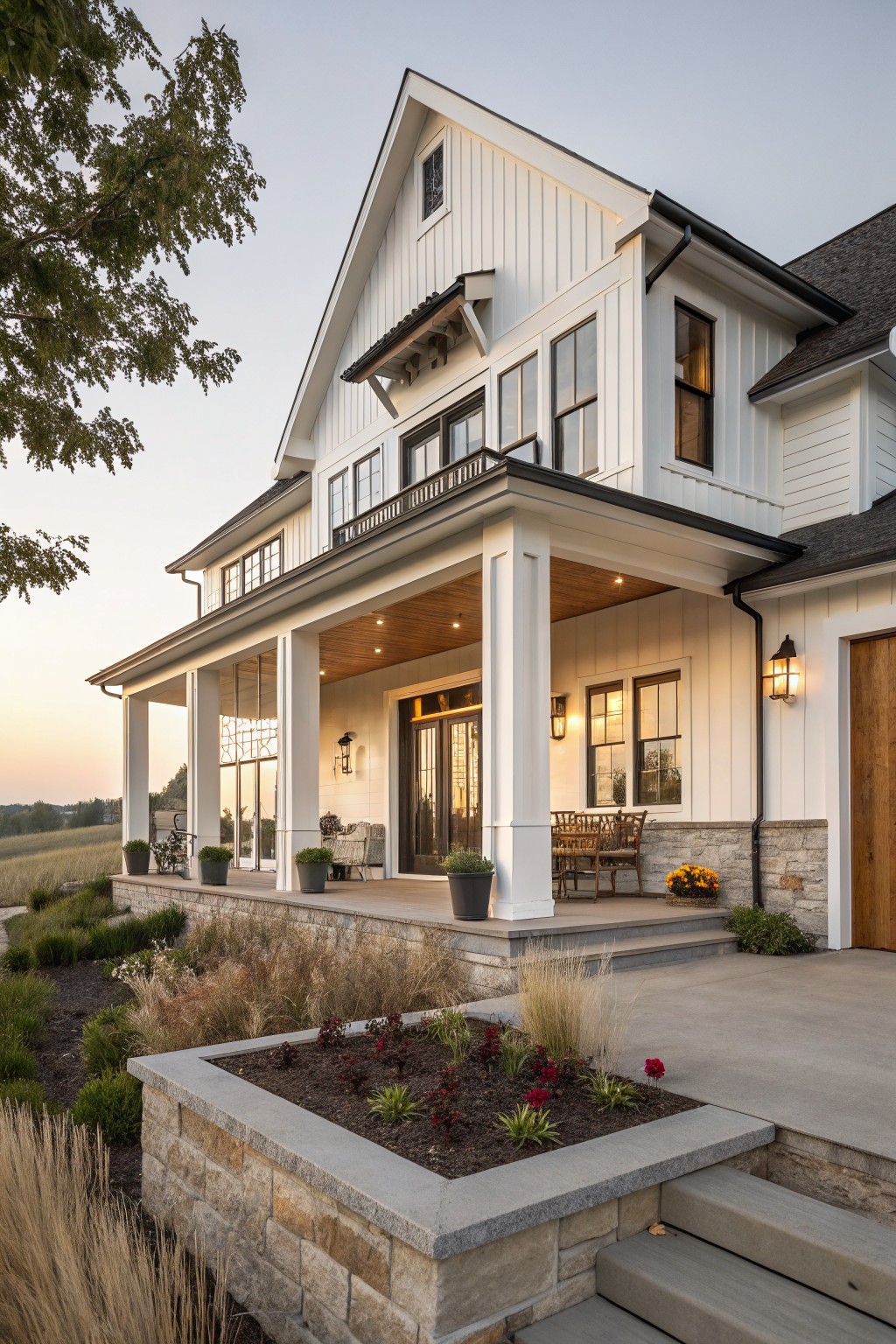 White board-and-batten sided modern farmhouse exterior with covered porch supported by tall white columns, double entry doors, stone retaining wall and steps with plants, and adjacent garage door at dusk.