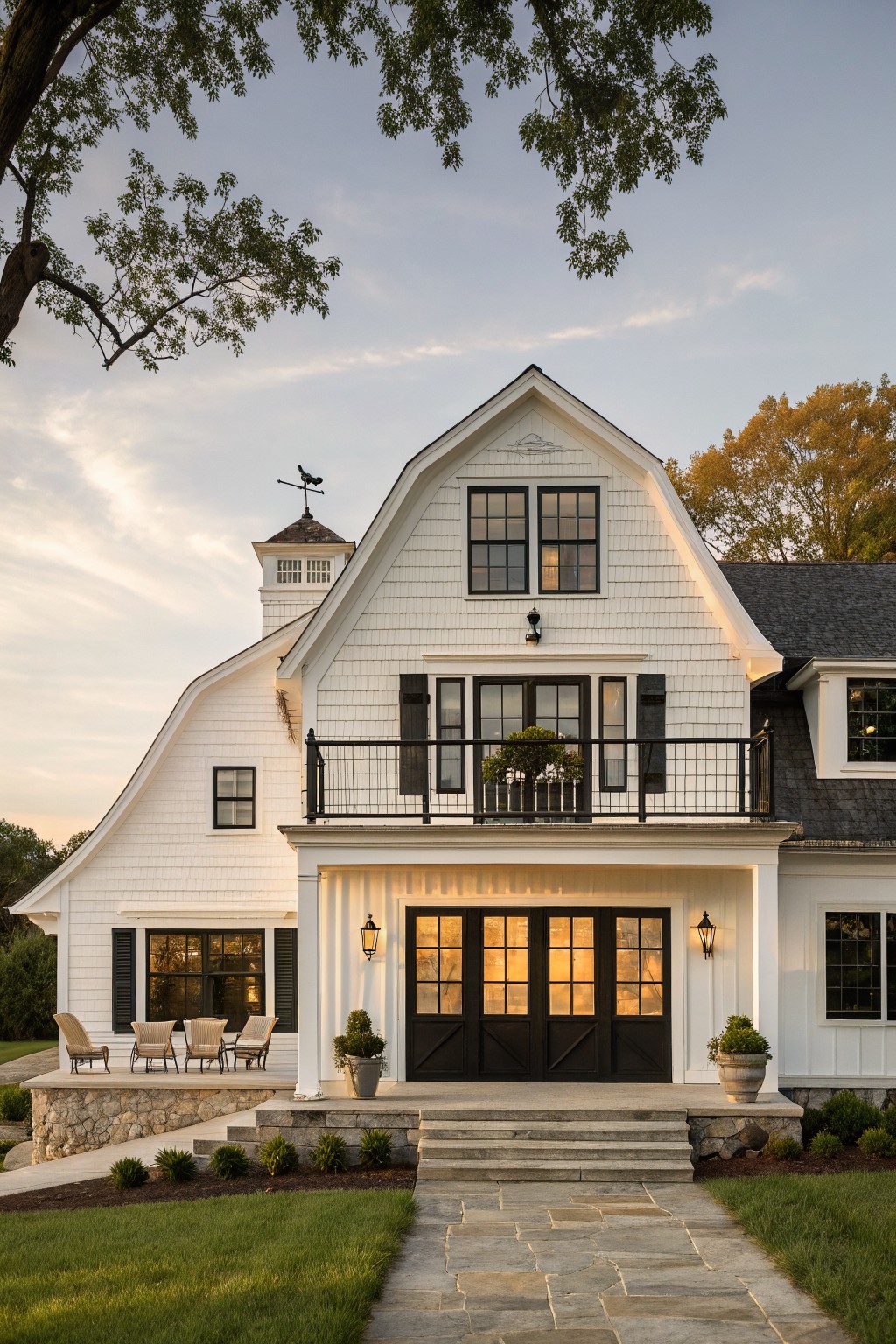 White shiplap farmhouse exterior with gambrel barn roof, cupola, black multi-pane front doors on columned porch, Adirondack chairs, lanterns, stone steps, bluestone path, shrubs, trees, and lawn at dusk.