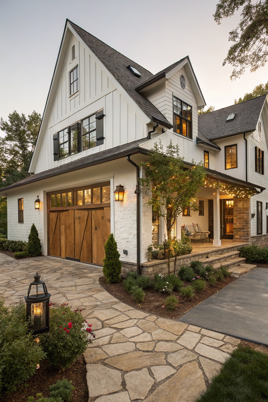 White two-story modern farmhouse exterior with attached garage featuring tall wooden carriage doors, covered porch with string lights, stone steps, flagstone pathway, shrubs, and lanterns at dusk.