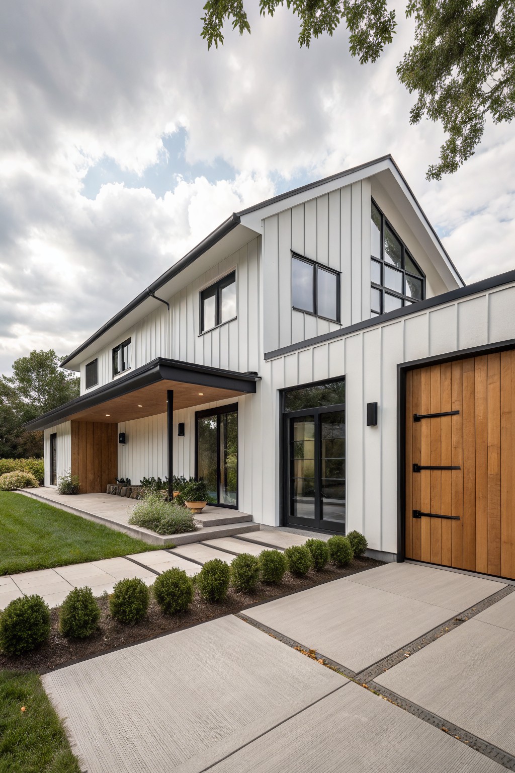 White modern farmhouse exterior with vertical board-and-batten siding, black-trimmed windows and roof edges, large wooden garage door with black hardware, covered entry porch, concrete walkway with boxwood shrubs, and surrounding lawn and trees under cloudy skies.
