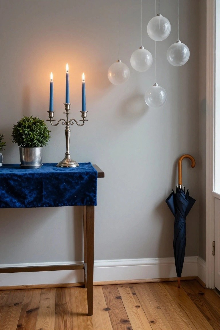 Gray corner of an interior room featuring a wooden console table with blue velvet runner, silver candelabra holding three lit blue candles, potted topiary plant in metal pot, multiple hanging clear glass sphere ornaments, navy umbrella leaning by window, and hardwood floors.