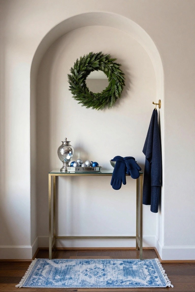 Arched white wall niche with gold console table holding silver cloche jar, blue and silver Christmas ornaments in a tray, navy knit gloves, evergreen wreath on wall, navy coat on gold hook, and blue patterned rug on wood floor.