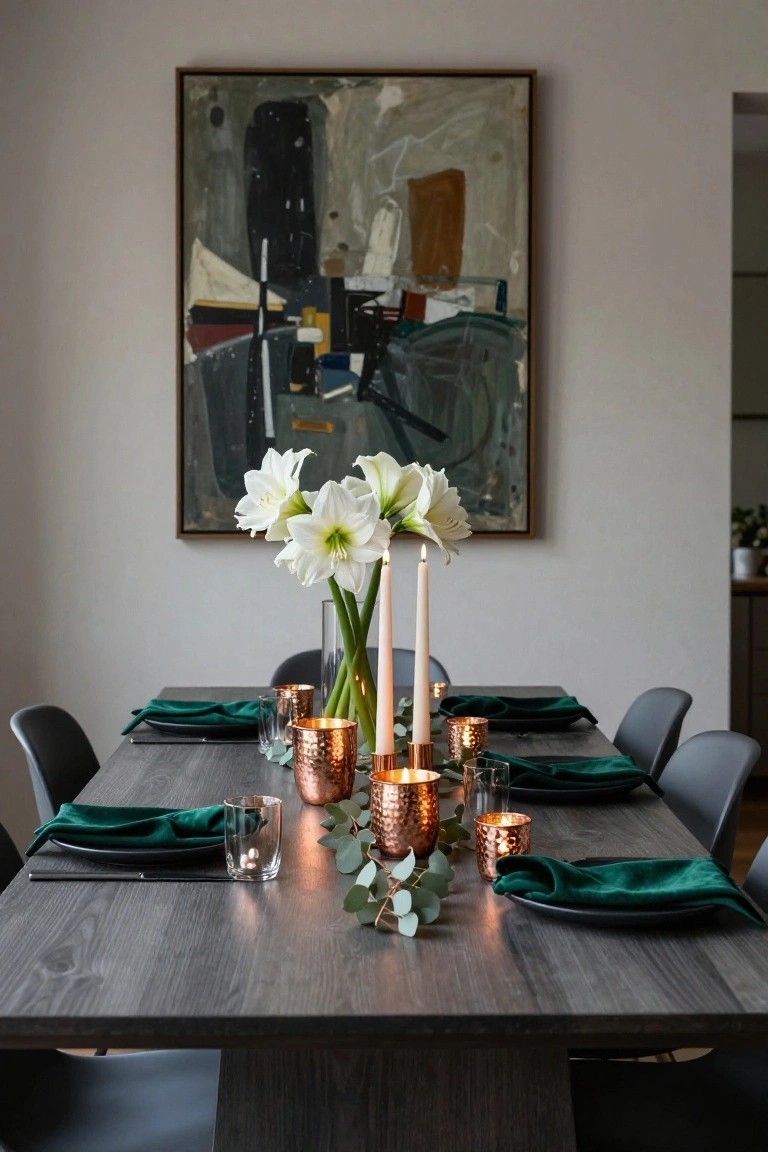 Dining table with green napkins and black plates, central tall vase of white lilies flanked by two taper candles, hammered copper cups holding tealights and eucalyptus leaves, grey chairs, and abstract painting on the wall.