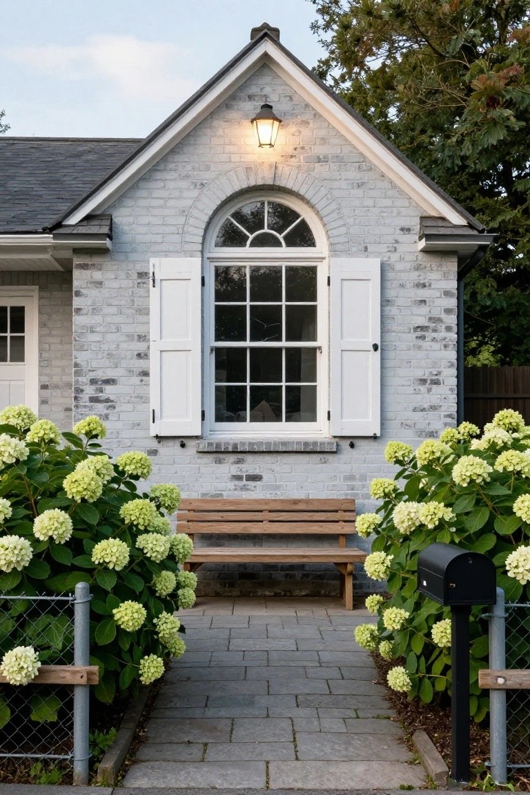 White brick cottage exterior with gabled roof, arched window with shutters, wall lantern, stone pathway, wooden bench, green hydrangea bushes, chain link fence, and black mailbox.