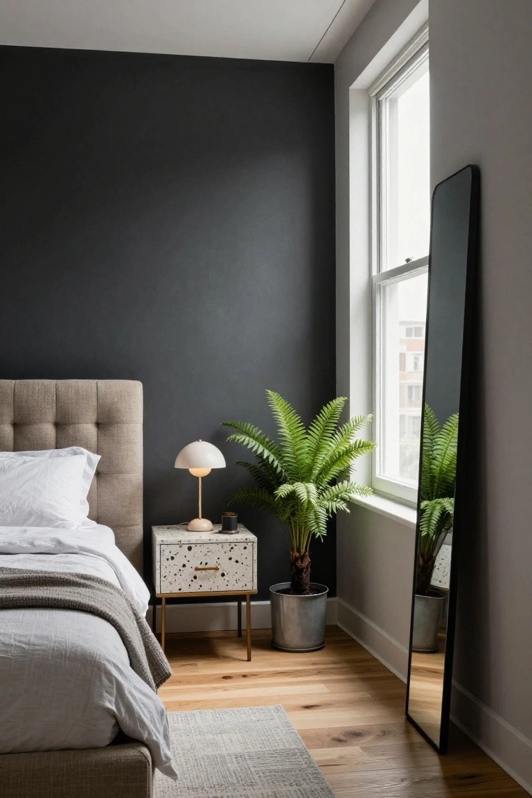 Bedroom with dark charcoal walls, beige tufted bed with white linens and gray throw, nightstand topped with beige lamp and candle, tall fern plant in metal pot, slim black floor mirror, hardwood floors, and large window.