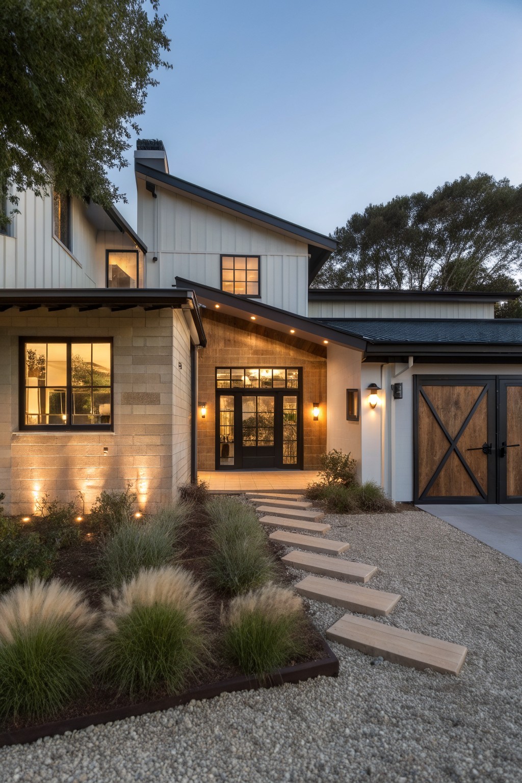 Modern ranch house exterior with white vertical siding, light beige stone base, double black X-pattern barn garage doors, wood-framed glass entry doors, pathway of wide wooden steps through gravel and ornamental grasses, surrounded by trees at dusk.