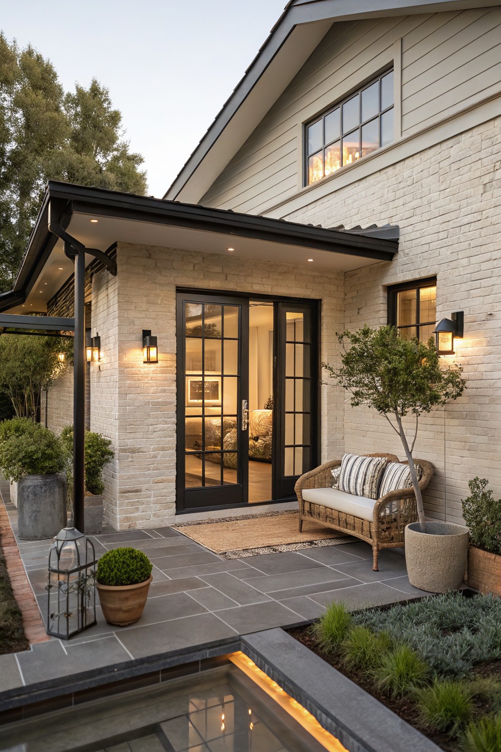 Side exterior of a ranch-style house with light beige brick walls, black metal-framed glass doors and windows, a covered porch area with lanterns, potted plants, wicker bench, and stone patio next to a pool edge.