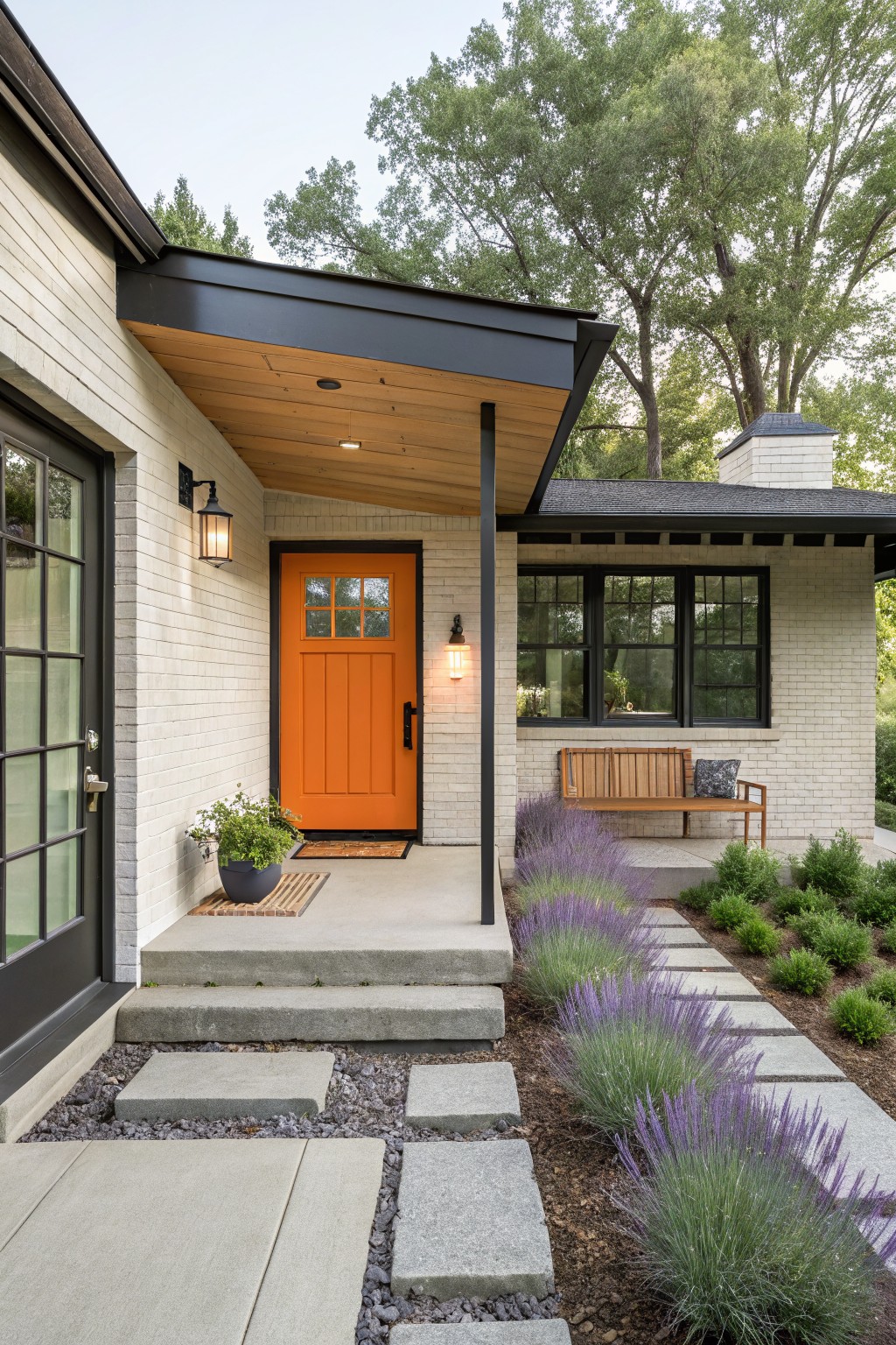 White brick ranch house exterior featuring an orange wood front door with glass panels, flanked by black-framed windows, a wood bench, lanterns, and purple lavender plants along concrete steps and path.