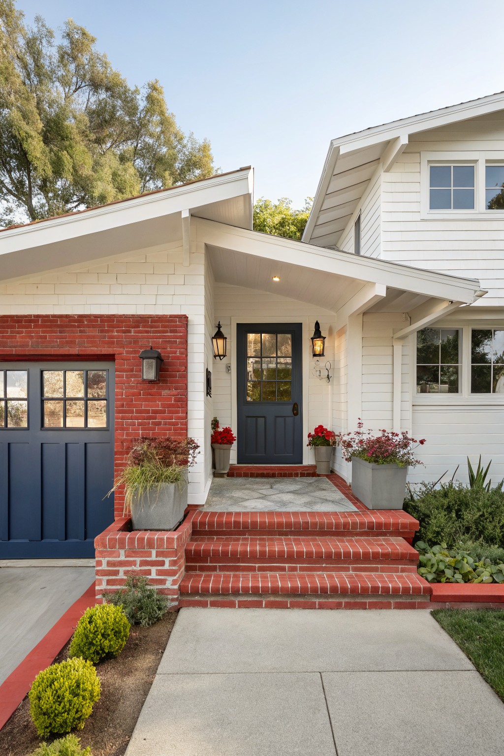 White ranch-style house facade with red brick base around navy blue paneled garage door and entry door, brick steps leading to the door, white siding above, lanterns, and potted plants on a concrete path.